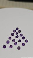 Photo of 22 round, faceted, calibrated amethyst gemstones arranged on a white surface showing the deep purple color and diamond cut facets.