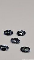 Five natural calibrated oval cut tanzanite gemstones placed on a plain surface showing color variations in blue, purple, and green tones