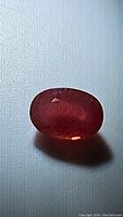 Single deep red-orange oval cut sapphire gemstone on a plain background, showing color and cut details