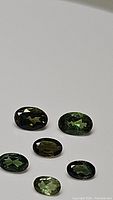 Front view of six oval cut green tourmaline gemstones with varying green shades on a white background.