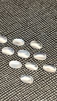 Close-up photo of 13 oval cabochon white moonstones laid out on textured black surface, showing translucence and catseye flash.