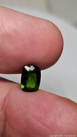 Close-up photo of the long cushion cut natural Chrome Diopside showing the deep electric green color and facets held between fingers.