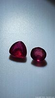 Two natural ruby gemstones laid on textured surface, showing one trillion cut and one oval cut stone in deep red color.