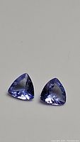 Photo showing two natural trillion cut tanzanite gemstones, purple blue with strong deep color, placed on grey background.