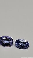 Two oval-cut purple-blue tanzanite gemstones placed side by side on a neutral background showing color and faceting.