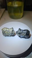 Two pig gemstone figurines, one dalmatian jasper and one darker stone, shown side by side on a round white surface.
