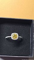 Single view of the ring in black jewelry box showing central yellow moissanite and surrounding white stones on band and setting.