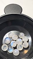 Top-down photo showing a group of 12 round moonstone cabochon gems in a black container, displaying a blue flash.