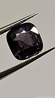 Close-up photo of a purple cushion cut spinel gemstone held by tweezers, showcasing facets and color under lighting conditions.