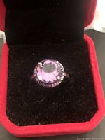 Front angled view of the ring showing the large faceted amethyst and surrounding smaller stones in a sterling silver setting inside a red and black jewelry box.