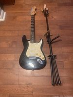 Full view of black Fender Squier Stratocaster electric guitar laying flat on wood floor next to black guitar stand