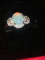 Close-up photo of opal ring showing oval cabochon gemstone and clustered clear accent stones on a polished sterling silver band resting on black velvet.