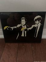 Framed street art print showing two men in white suits holding yellow bananas against a black background, viewed straight on.