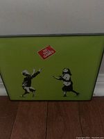 Framed street art print showing a green background with two children reaching up towards a red 'No Ball Games' sign.