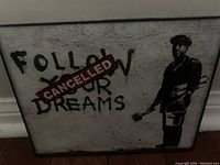 Framed street art print with stencil image of man and text 'FOLLOW YOUR DREAMS', with 'CANCELLED' stamped over 'YOUR'. The print appears to be behind glass with a black frame.