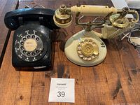 Two antique rotary dial telephones, one black and one cream-colored with gold accents, placed on a wooden surface.