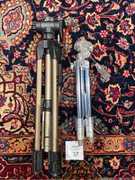 Two camera tripods, one gold and black, one silver with transparent mount, laid on a patterned carpet
