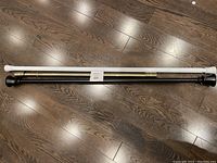 Two extendable curtain rods placed side by side on a wooden floor, one black with gold accents and one white, with no mounting hardware shown.