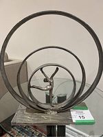Metal sculpture featuring a dancer performing inside three concentric circular metal rings on a rectangular base, photographed indoors on a glass table.
