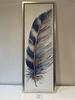 Photo of framed feather print hanging on a light colored wall showing the full artwork vertically, highlighting blue, green, and grey tones.