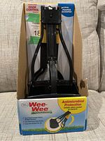 Front view of the fold-away pooper scooper partially assembled in its original cardboard box, showing the black plastic scoop and folding frame.