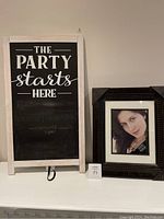 Photo showing a white easel holding a chalkboard sign with the text 'The Party starts here' and a separate black textured photo frame with an image of a woman.