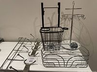 Five metal hanging baskets and wall hangers displayed on floor and against wall, including jewelry holder, blow dryer basket, suction cup shower basket, and two hanger racks.