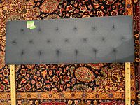 Full view of dark blue fabric upholstered headboard showing tufted button design and wooden legs on a patterned rug.