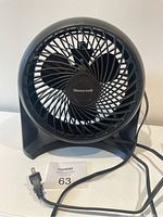 Front view of black Honeywell plug-in fan with visible blades and Honeywell logo in center, power cord and plug placed in front of fan.