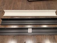 Three Ikea Mosslanda wall shelves on a wooden floor: two black and one white, approximately 115 cm long each, no mounting hardware.