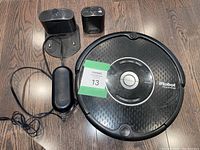 Top view of black iRobot Roomba vacuum robot, charging base, remote control, and small accessory on wooden floor.