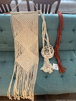 Three macrame pieces including a large wall hanging and two plant holders on a teal couch.