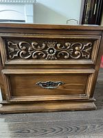 Front view of the wooden night stand showing two drawers. The upper drawer has intricate carved floral design and metal pull, the lower drawer has an ornate metal handle.