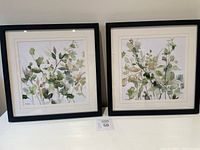 Pair of framed botanical watercolor prints with leafy green and beige designs, set against white mats and black frames.