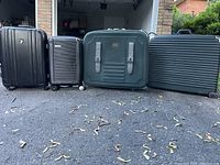 Four suitcases in a row on driveway; one is green with straps, others are black with ridged surfaces; all have wheels.