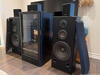 Pair of large Technics floor-standing speakers with removable grills leaning against them and black wood-finished audio component cabinet in between.