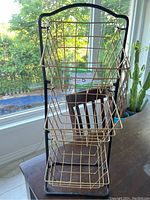 Photo showing a three tier wire basket organizer with three gold metal baskets stacked vertically on a black metal frame near a window.