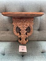 Front view of decorative wooden wall shelf showing thickness of the shelf top and ornate carved bracket with floral and scroll details.