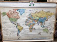 Photo of large rectangular cork board with world map glued on it, framed with white wood, showing continents, countries, oceans, and political borders in color.