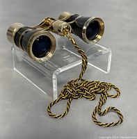 Front angled view of black and gold opera glasses on clear stand showing the eyepieces and attached gold and black chain coiled in front.