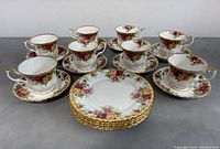 8 floral Royal Albert teacups each with matching saucers, and 5 floral side plates, all decorated with pink and yellow roses bordered with gold trim, arranged on a grey surface.