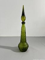 Front view of a tall green green pressed glass Mid-Century Modern style decanter with embossed decorative patterns and a long pointed stopper, set against a plain backdrop.