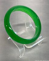 Close-up photo of the polished bright green jade bangle on a clear display stand, showing smooth surface and shape.