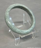 Natural jade bangle bracelet on clear stand, showing rounded polished surface and color variation.