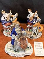 Front view of the cellist figurine wearing blue coat, holding cello on ornate base with sheet music and floral accents