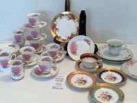 Overall arrangement showing multiple teacups and saucers on a white surface with floral patterns in pink and gold accents, including two dark blue rimmed plates with floral motifs.