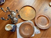 Overview of lot items displaying two silver-plated candelabras, a brass tray, two copper plates, and a silver posy bowl on a wooden floor.