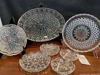 Wide shot showing 2 platters (oval and round) standing upright on clear and black stands and 5 smaller crystal bowls arranged in front on a wooden surface.