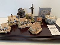 Collection of six Lilliput Lane cottages alongside Japanese bowl, metal statue, Alaska decor piece on dark wooden surface.