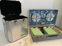 Photo showing the picnic set case with blue lining holding cutlery, plates with floral pattern, salt and pepper shakers, two green plastic containers, and four clear glasses, next to a closed silver aluminum storage case.